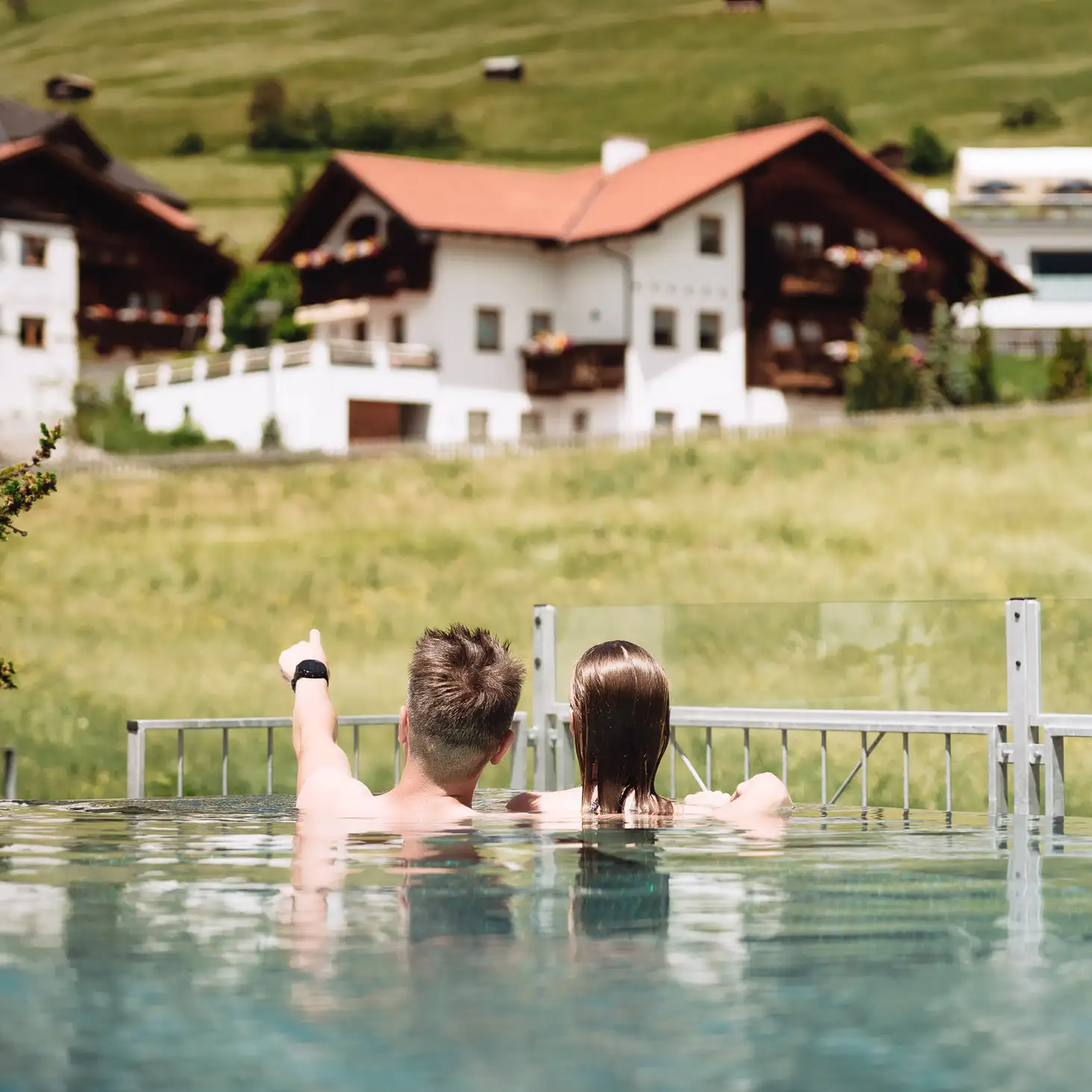 Zwei Personen in einem Pool blicken auf Alpenhäuser auf einem grasbewachsenen Hügel im Hintergrund.