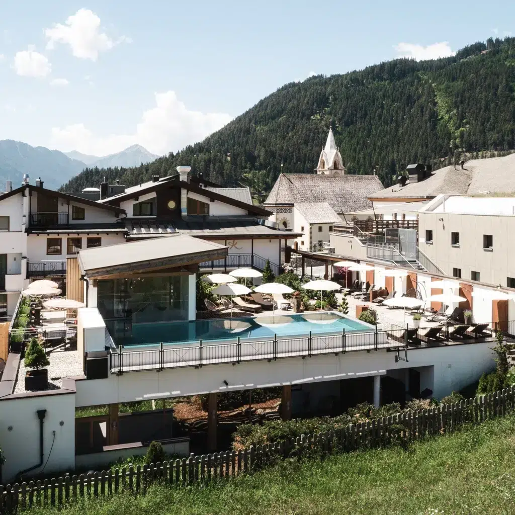 Modernes Hotel Das Köhle mit Pool auf dem Dach, Blick auf die Berge und traditionelle Gebäude an einem sonnigen Tag.