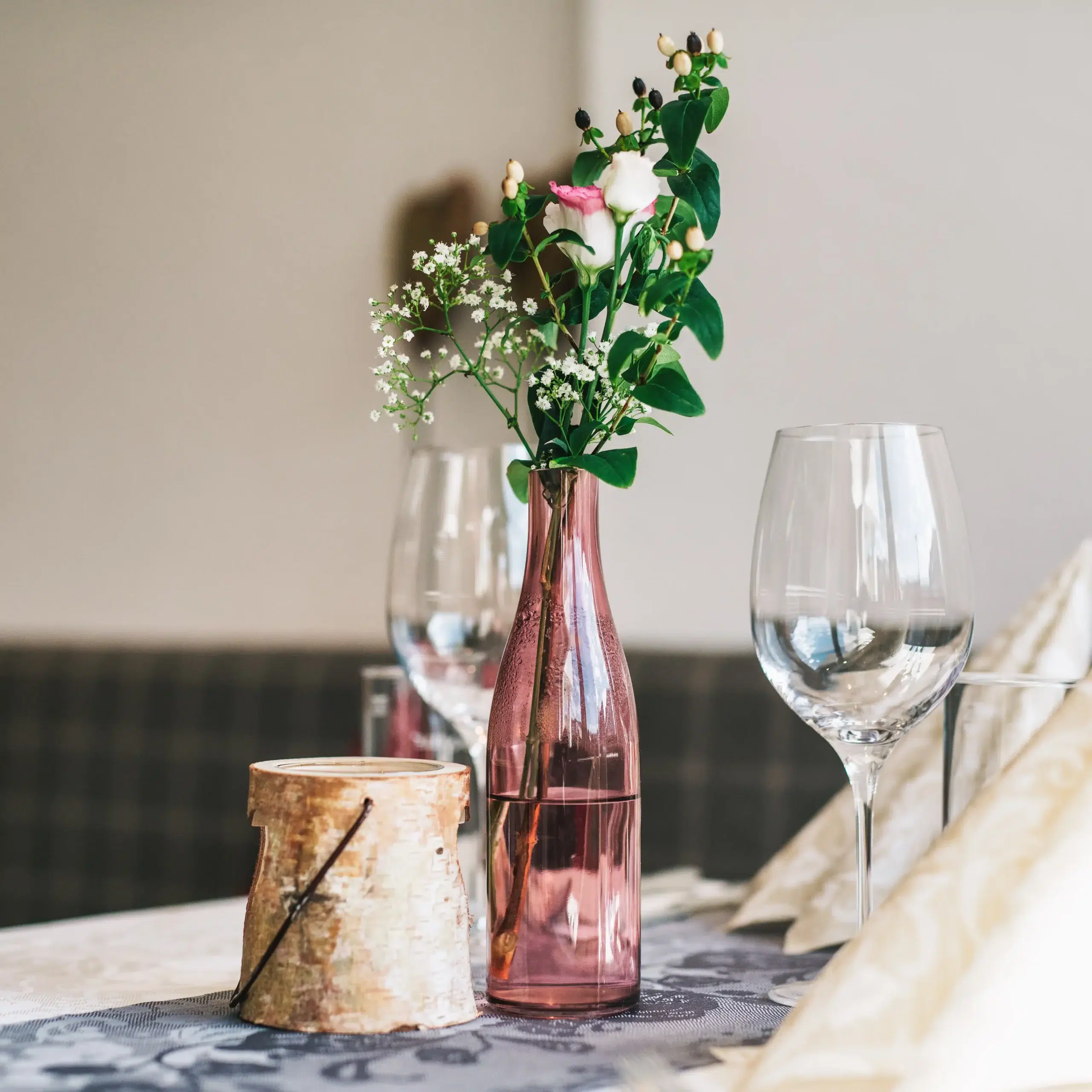 Rosa Glasvase mit Blumen, Weingläsern, und FAQ & Buchungsinfos auf einem Tisch.