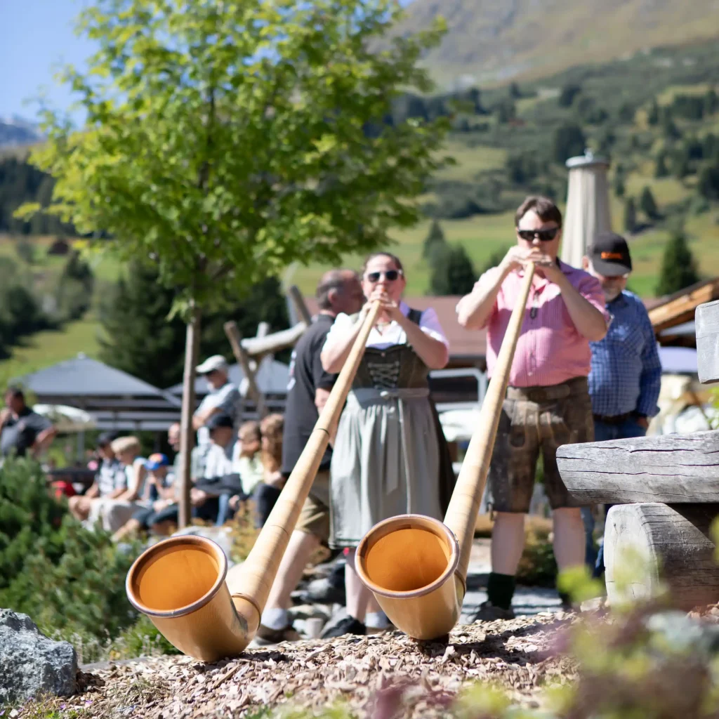 Zwei Personen spielen Alphörner im Freien beim Golden Fine Arts Festival mit den Bergen im Hintergrund.