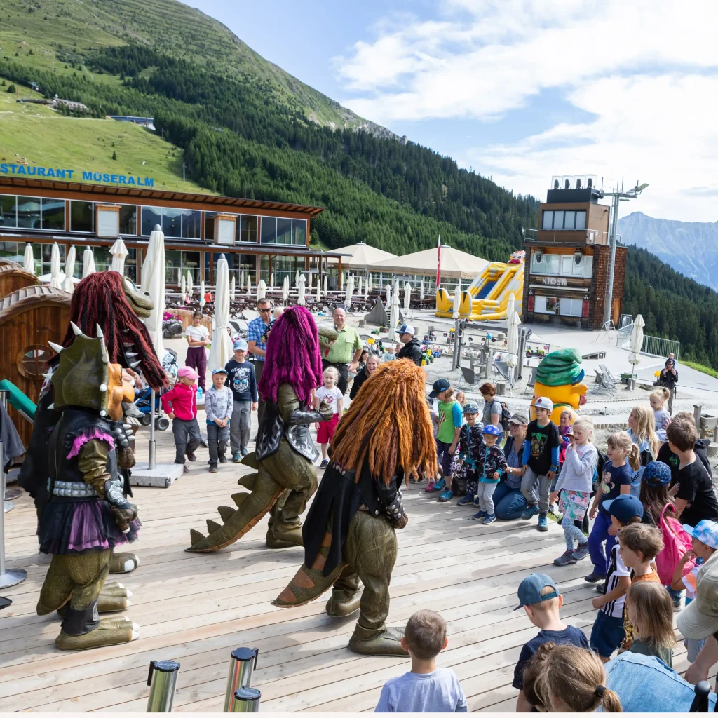 Menschen und Kinder beobachten den Auftritt kostümierter Maskottchen auf einem Holzdeck inmitten von Berghügeln.